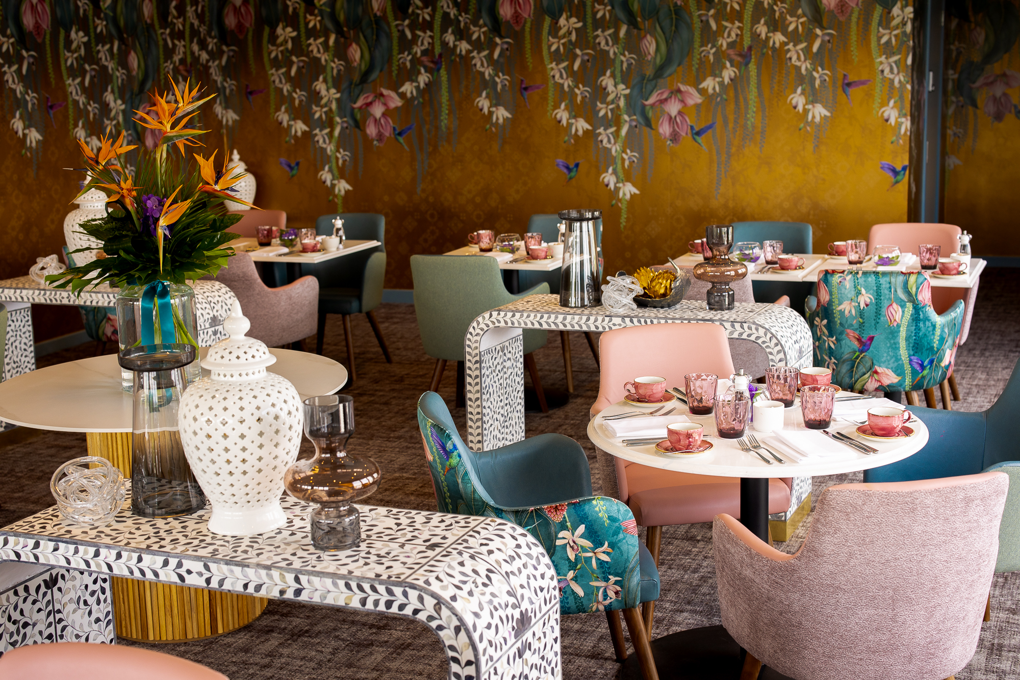 A dining room with multiple small tables set with tea cups and glasses, featuring colorful chairs and a wall with floral and hummingbird patterns.