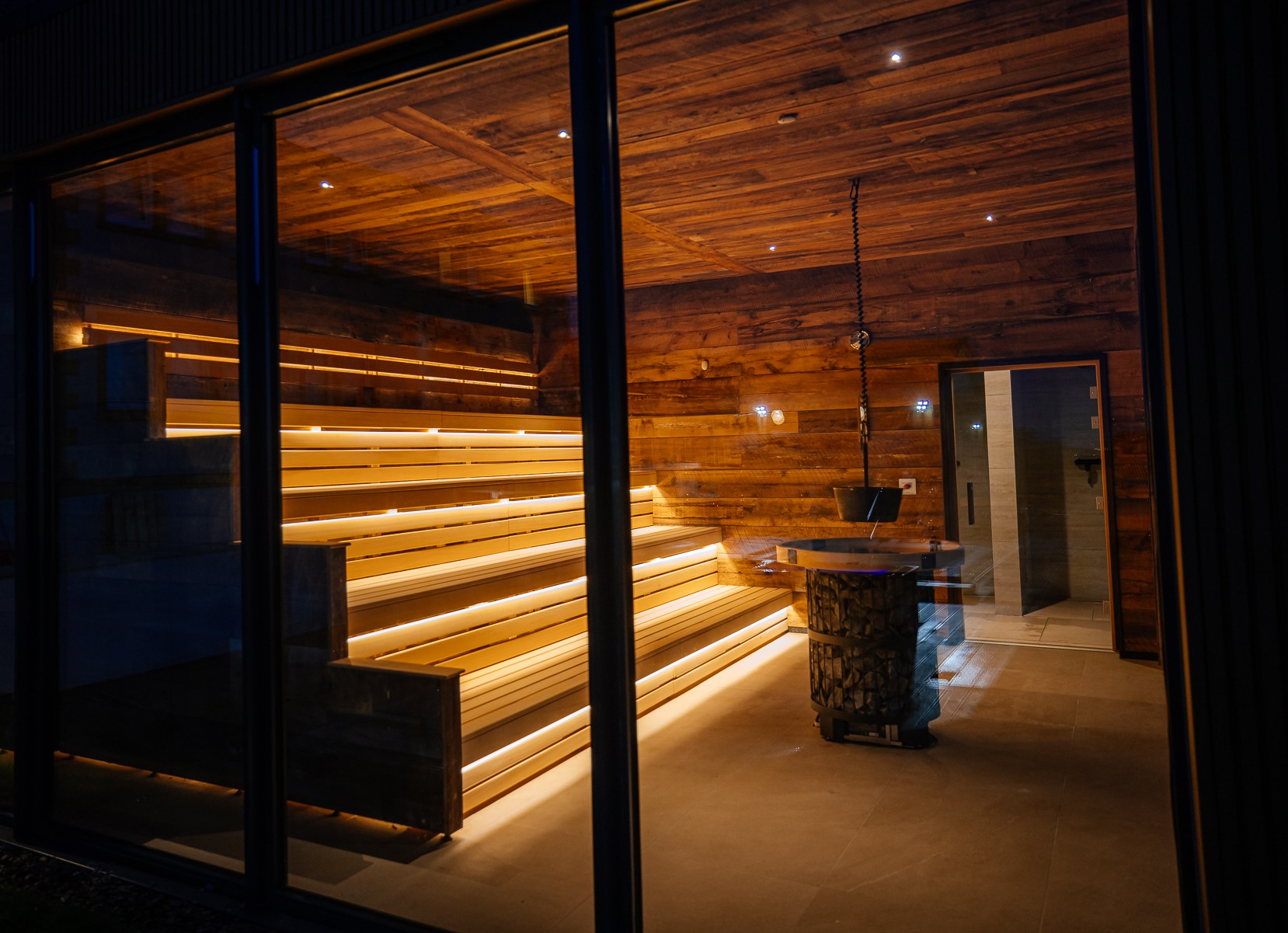 A wooden sauna with tiered seating, illuminated with warm lighting, and a metal sauna stove.