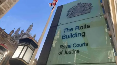Brian Farmer/BBC A white sign with the words "The Rolls Building Royal Courts of Justice" written on it in back. The to left of the sign is a brown building and a flag pole with a red, white and blue flag hanging loose at the top. Reflections of buildings can be seen in the sign.