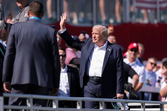 Person in a suit waves to a crowd at an outdoor event with spectators in the background