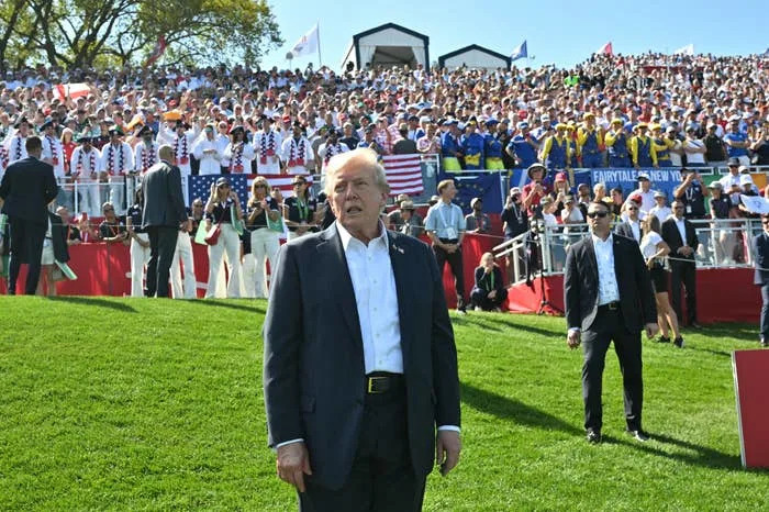 A man in a suit stands on a golf course with a large crowd in the background watching a golf event