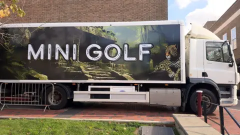 The Professional Idiots A lorry with mini golf on the side parked outside the townhall