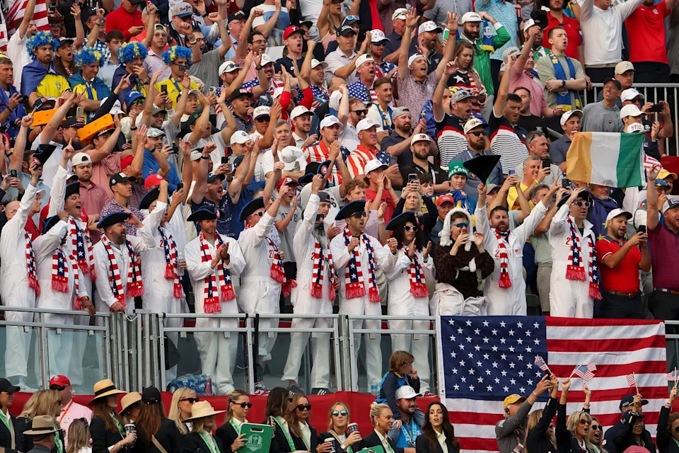 Crowd of diverse sports fans enthusiastically cheering at a stadium, some wearing white outfits and waving an American flag