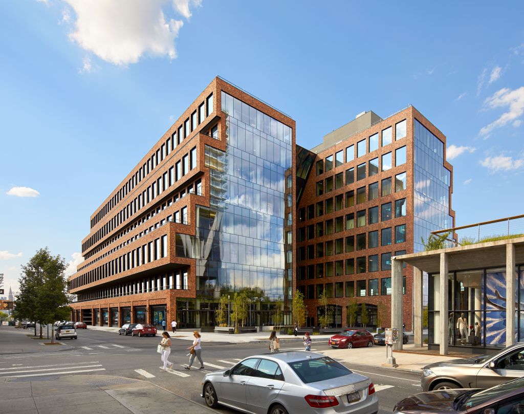 A large brick and glass building with several people walking on the sidewalk and cars parked along the street.
