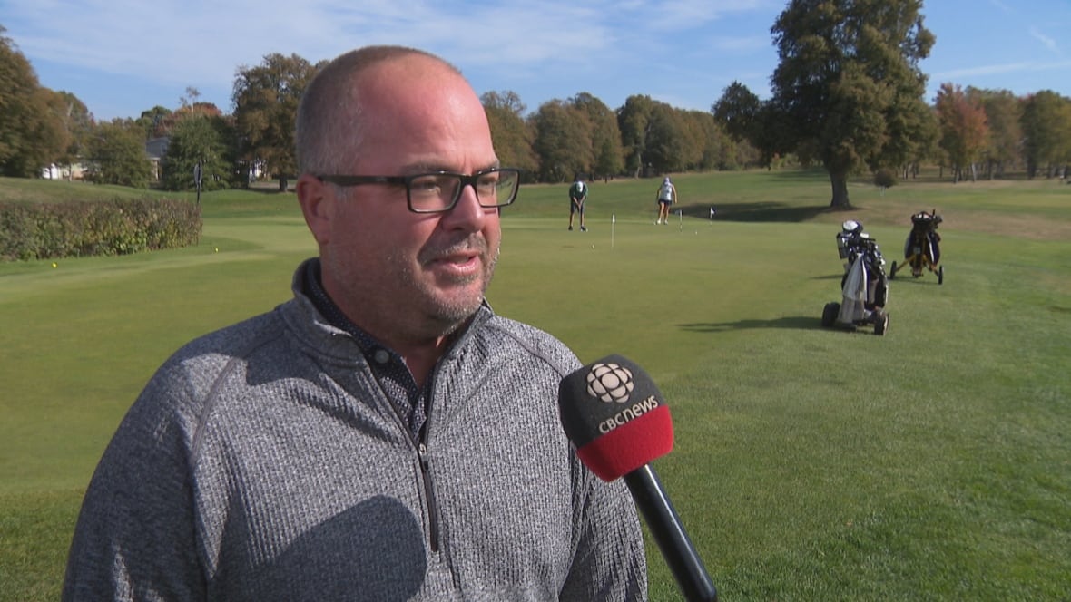 A man wearing glasses and a grey sweater stands in front of a golf course with golfers on it.
