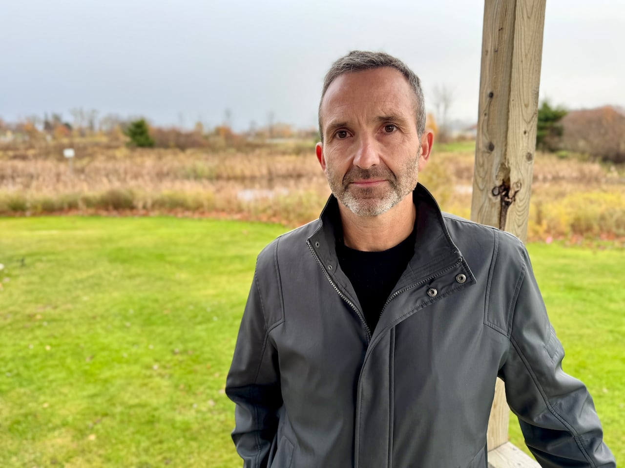 A man in a dark spring coat stands in font of a field. He is looking at the camera. 