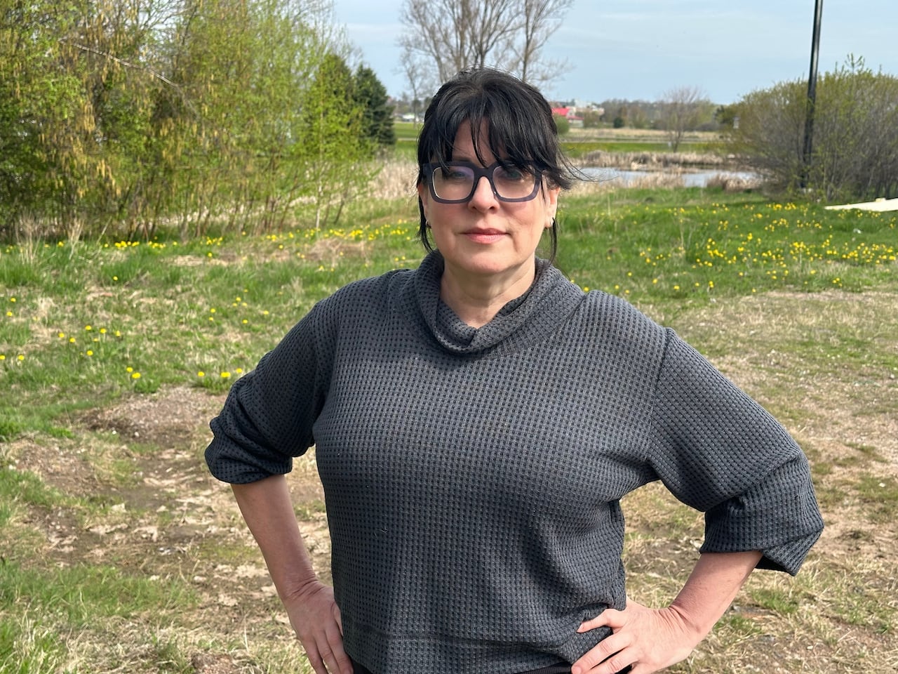 A woman with black hair and glasses, wearing a grey shirt, stands in front of a wetland. 