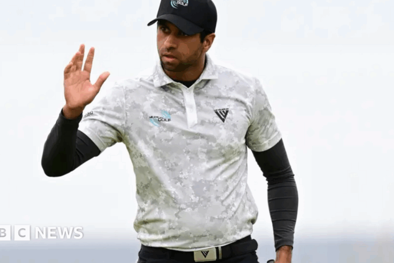Rai is wearing golf gear and raising his right hand as a wave possibly to spectators. He has a white top on over a long sleeve black top and a white cap.