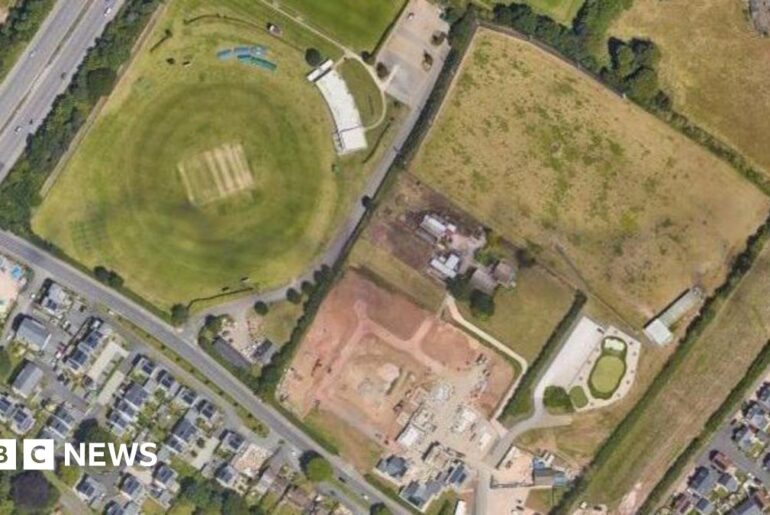 An aerial view of two fields which were formerly a golf driving range, surrounded by housing estates, roads and trees.