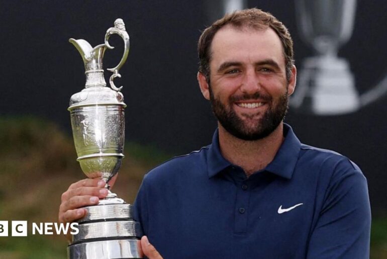 A man in a navy blue polo shirt, white trousers and a silver wrist watch holds up a trophy up and smiles at the camera. Behind him is a putting green and a sign which says "153rd Royal Portrush".