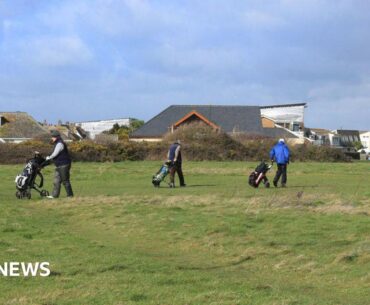 Three golfers with golf carts walking on the gold course at Solent Meads. In the background are neighbouring houses - large dormer bungalows with pitched roofs and two-storey modern houses with balconies.