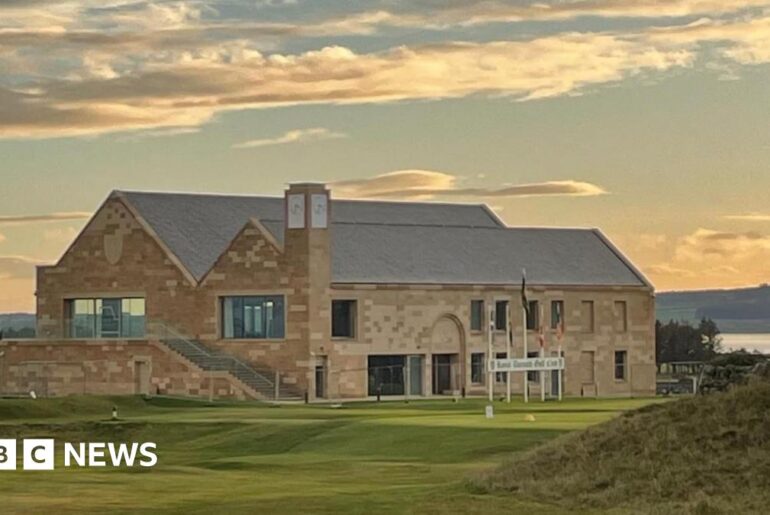 The sun has just gone down and clouds are lit in orange and gold colours above the new clubhouse at Royal Dornoch. It is a large building made of sandstone with a slate roof. A golf tee point is located in front of the building.