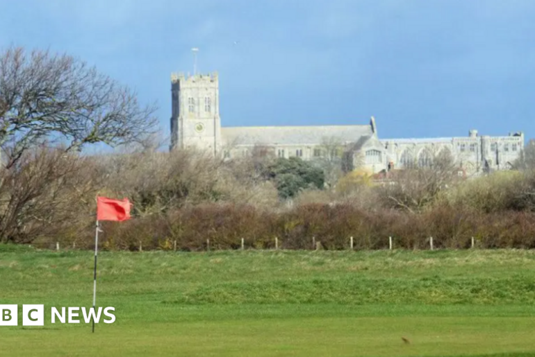 A telephoto shot of Christchurch Priory in the distance, with a putting green and an orange flag of Solent Meads gold course in the foreground. The priory church is a huge stone medieval building with a square tower at one end.