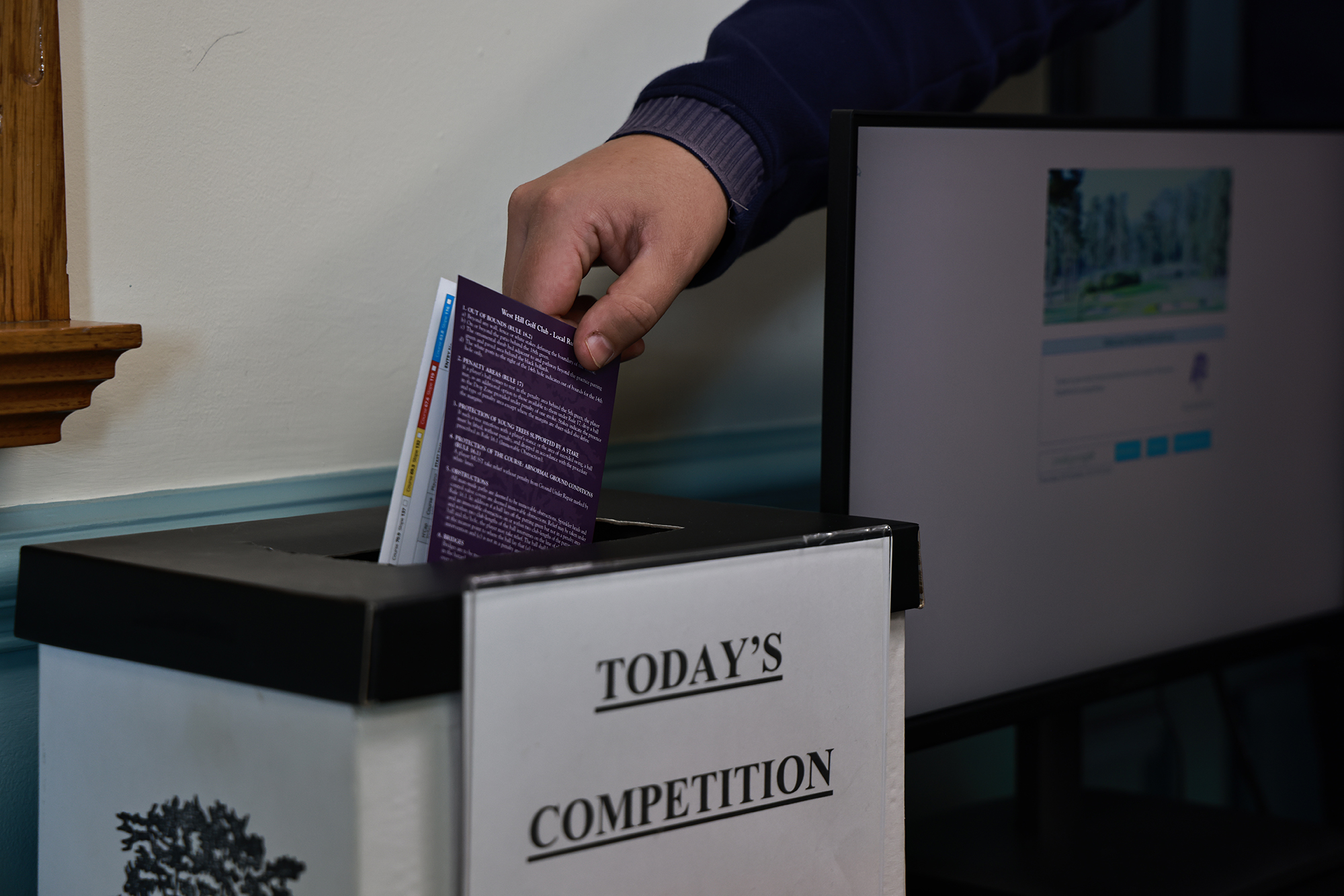 A golfer placing their scorecard into the box in the clubhouse that is labelled 'today's competitions', submitting their score in the process.
