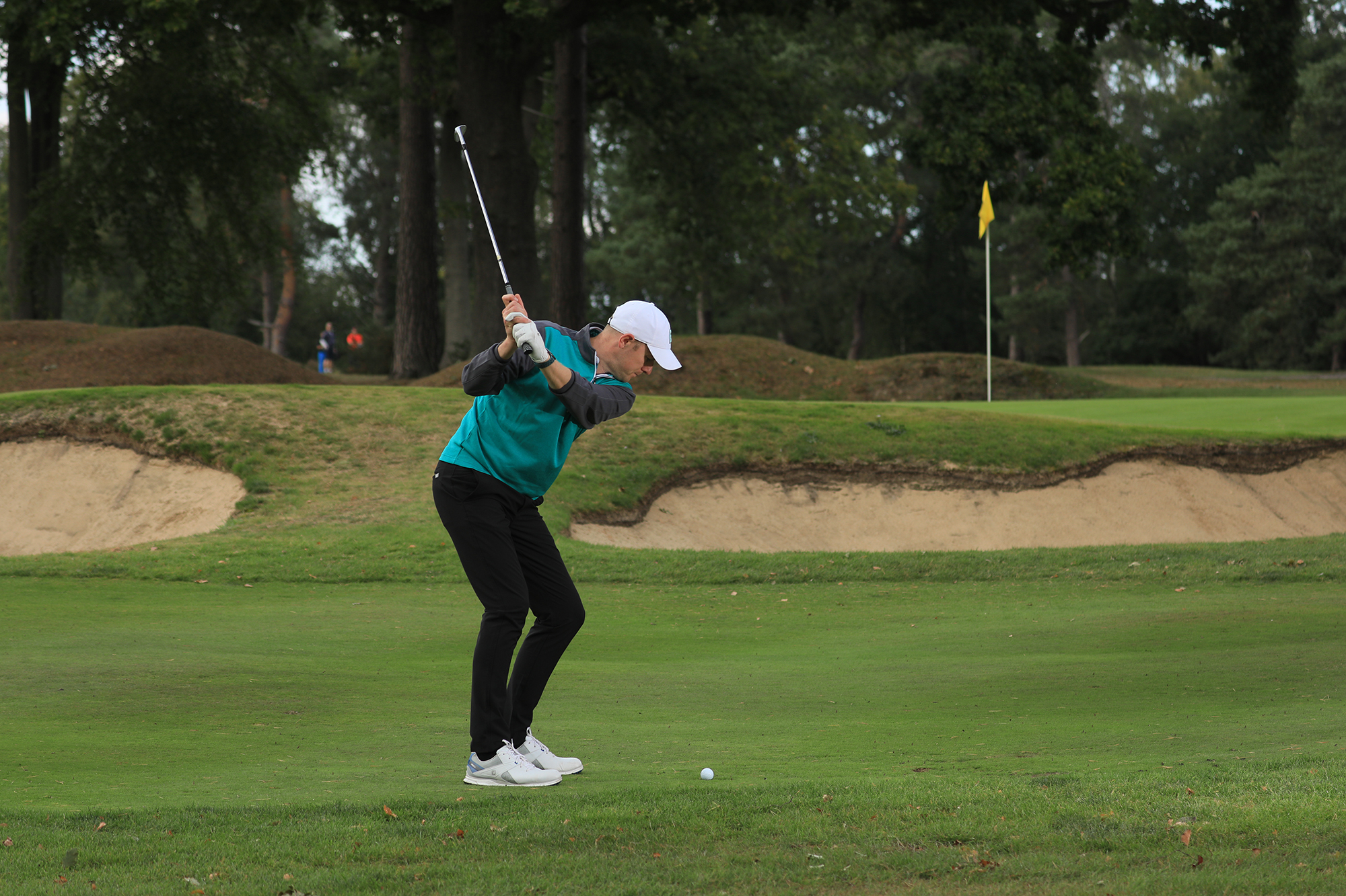 Neil Tappin hitting a pitch shot with a pitching wedge over a bunker towards a middle flag on the green