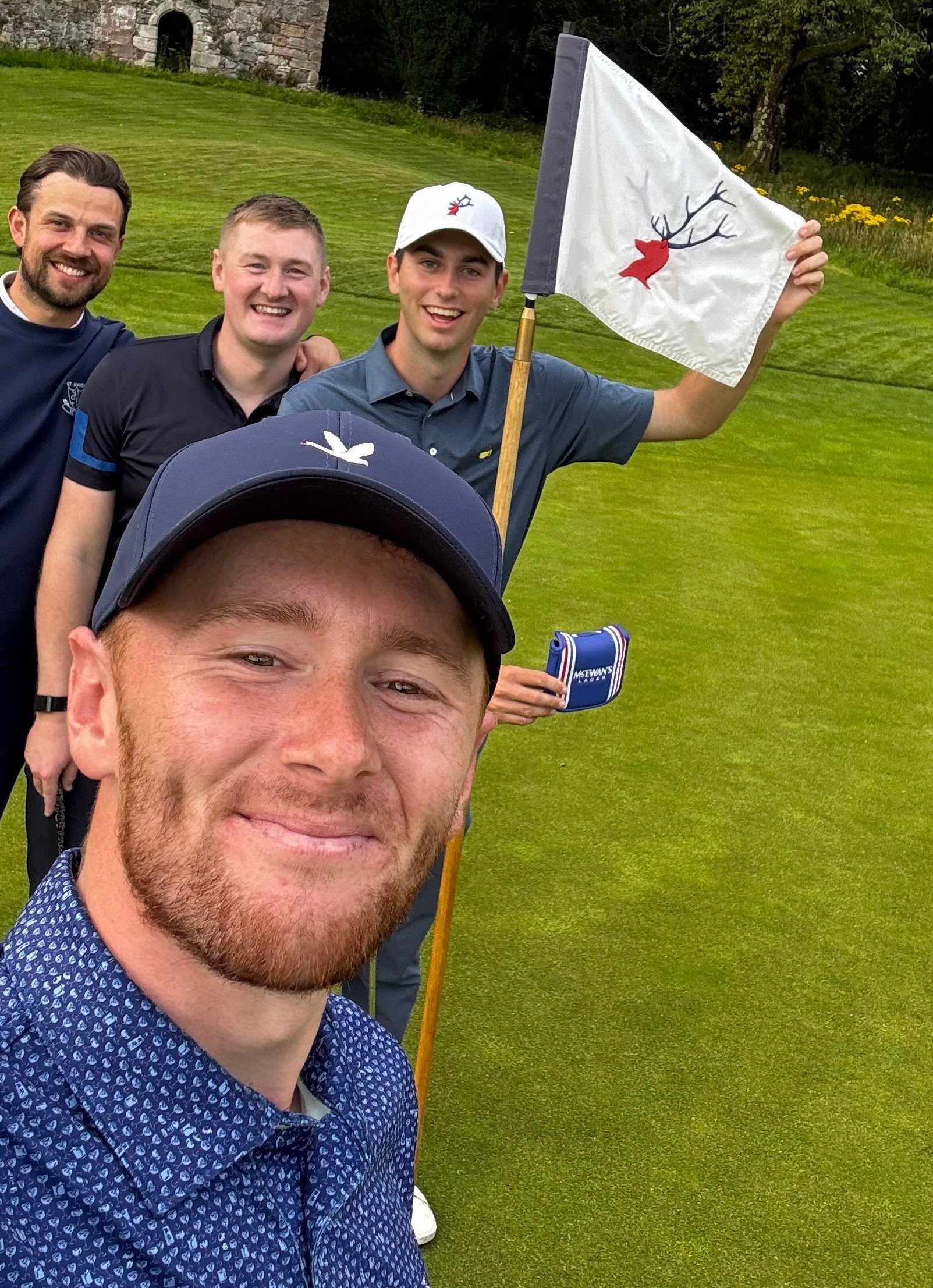 Josh Simpson, a red-bearded golfer, taking a selfie with three other golfers on a green, with an old stone building in the background.