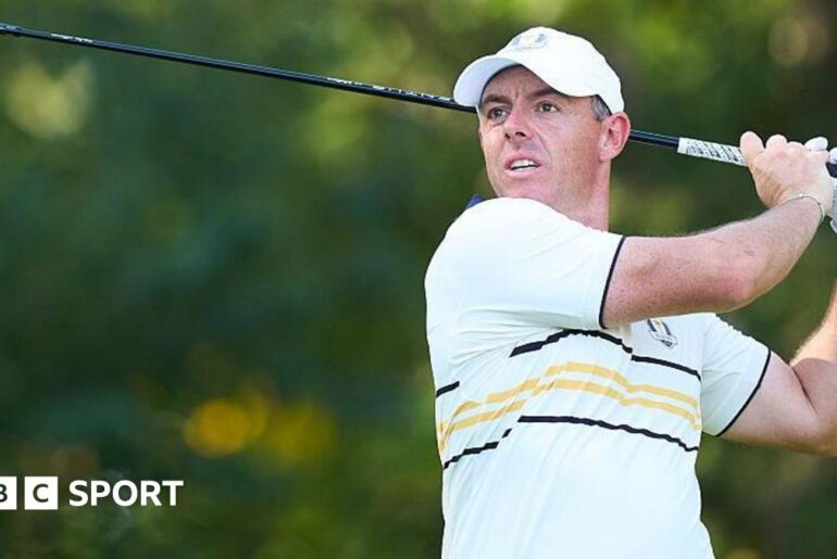 Rory McIlroy watches his ball after hitting a shot with a wood. He is wearing a European Ryder Cup outfit - a baseball cap and a predominantly white polo shirt with black and yellow narrow hoops and trim.