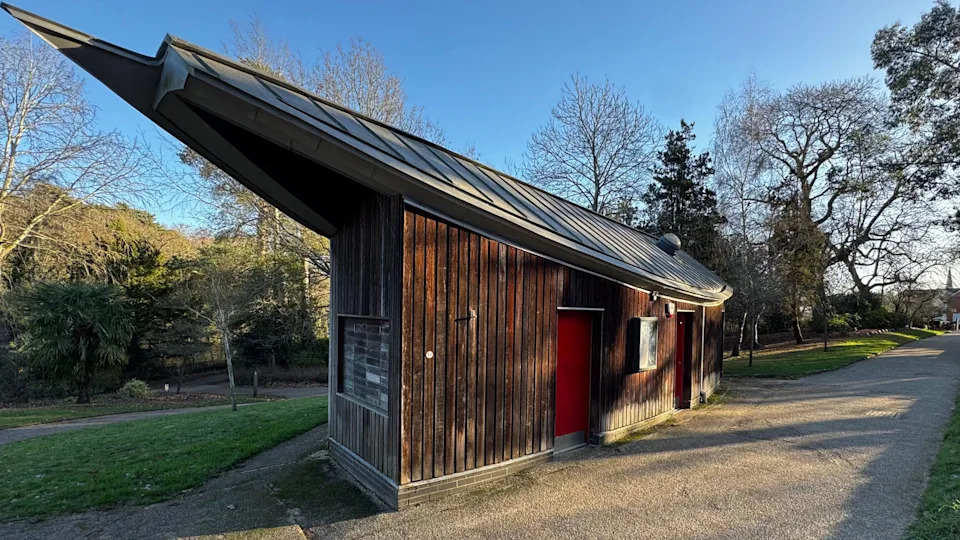 A large wooden hut with red doors and a pointy roof. Woodland and a concrete path surrounds it.