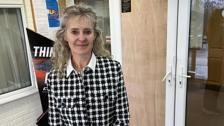 Director of golf at the West Malling club Kate Brett stands in front of a boarded-up door. She is wearing a black and white top with a white collar and has long, wavy blonde hair. There are club signs and posters behind her.