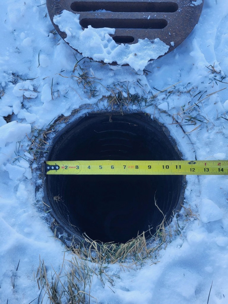 A measuring tape stretches across a dark, round pipe opening in the snow, next to an open manhole cover.