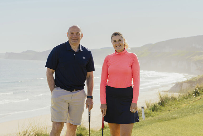 A man and woman standing on a golf course with a beach and mountainous background behind them.