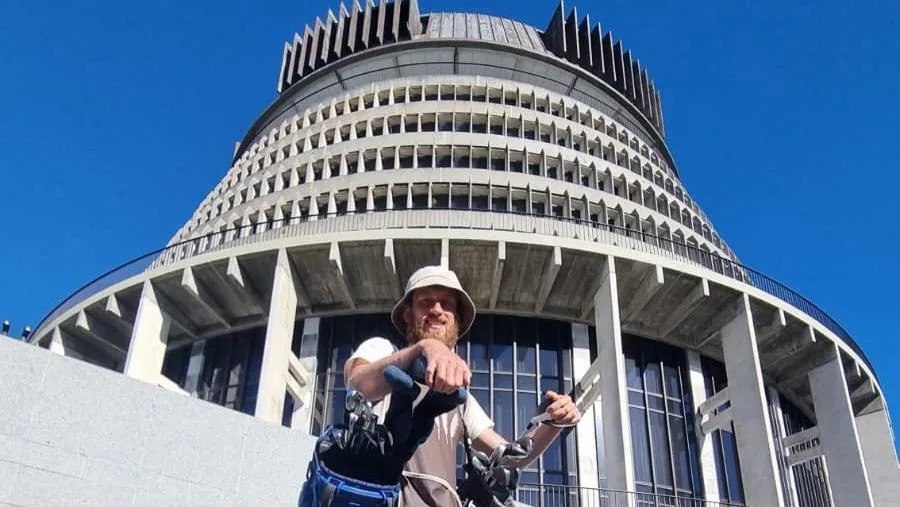 Dougie Haynes carrying two golf bags is standing in front of The Beehive, New Zealand's parliament building.