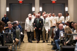 Group of men in white suit coats guide a man in a dark suit up an aisle.