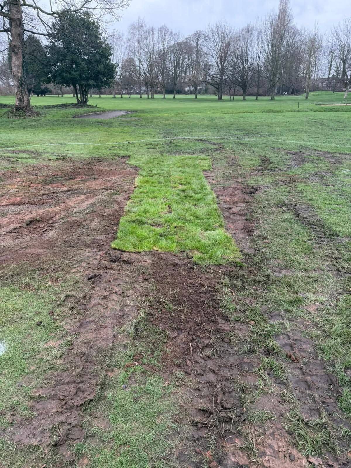 An outdoor scene depicting a section of a golf course undergoing landscaping. The field exhibits a mixture of bare earth and newly planted grass, indicating recent maintenance work. In the background, a landscape of trees and greenery is visible against a clear sky.