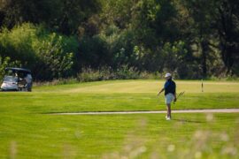 A men's golfer wearing a white hat and blue-striped polo follows through on his tee shot.