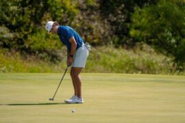 A men's golfer in a white hat and blue polo lines up his ball before attempting his put.