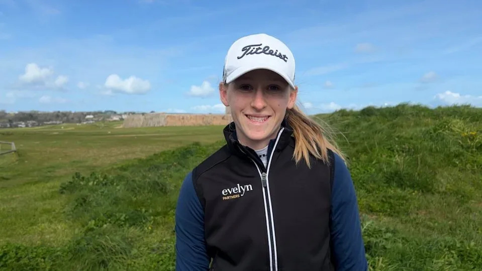 A young woman with light brown hair smiles into the camera. A white golf cap shadows her eyes. She's also wearing a black gilet over a dark blue sports jumper. Behind her is a golf course, with a green mound and old fortifications.