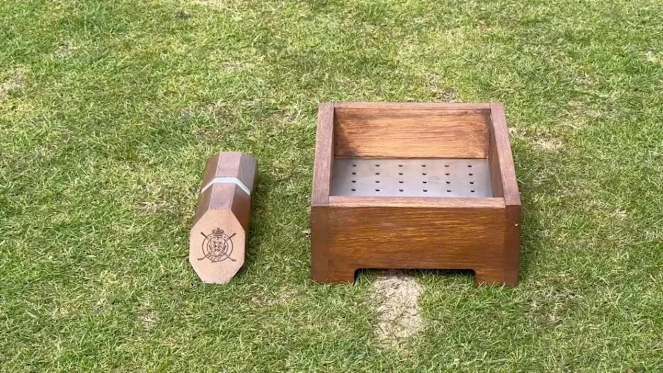 A wooden golf tee box marker, with a silver stripe down the middle sits on grass. On its right, an open-top wooden box contains a metal sheet with several holes.