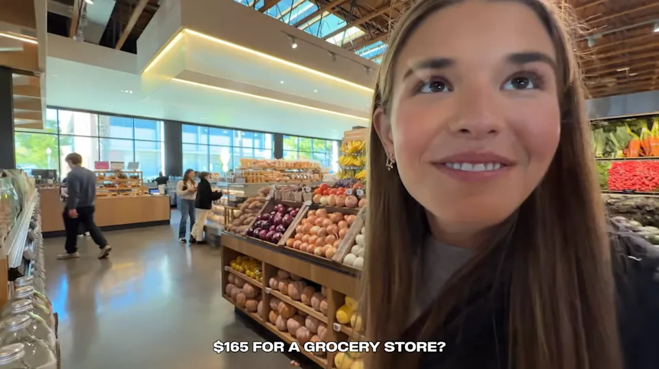 18-year-old woman grinning up at the camera in a luxury grocery store in the produce aisle saying, "$165 for a grocery store?"