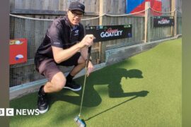 A man in all black squatting by the side of a mini golf course. He is holding a putter.