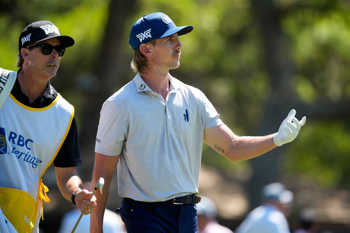 Two individuals are on a golf course, one wearing a white polo shirt and blue pants, the other donning a white and yellow vest and a black cap. Both are equipped with golf gloves, and the individual in white appears to be gesturing while the other looks on. The background features trees and other people, some of whom are wearing hats.