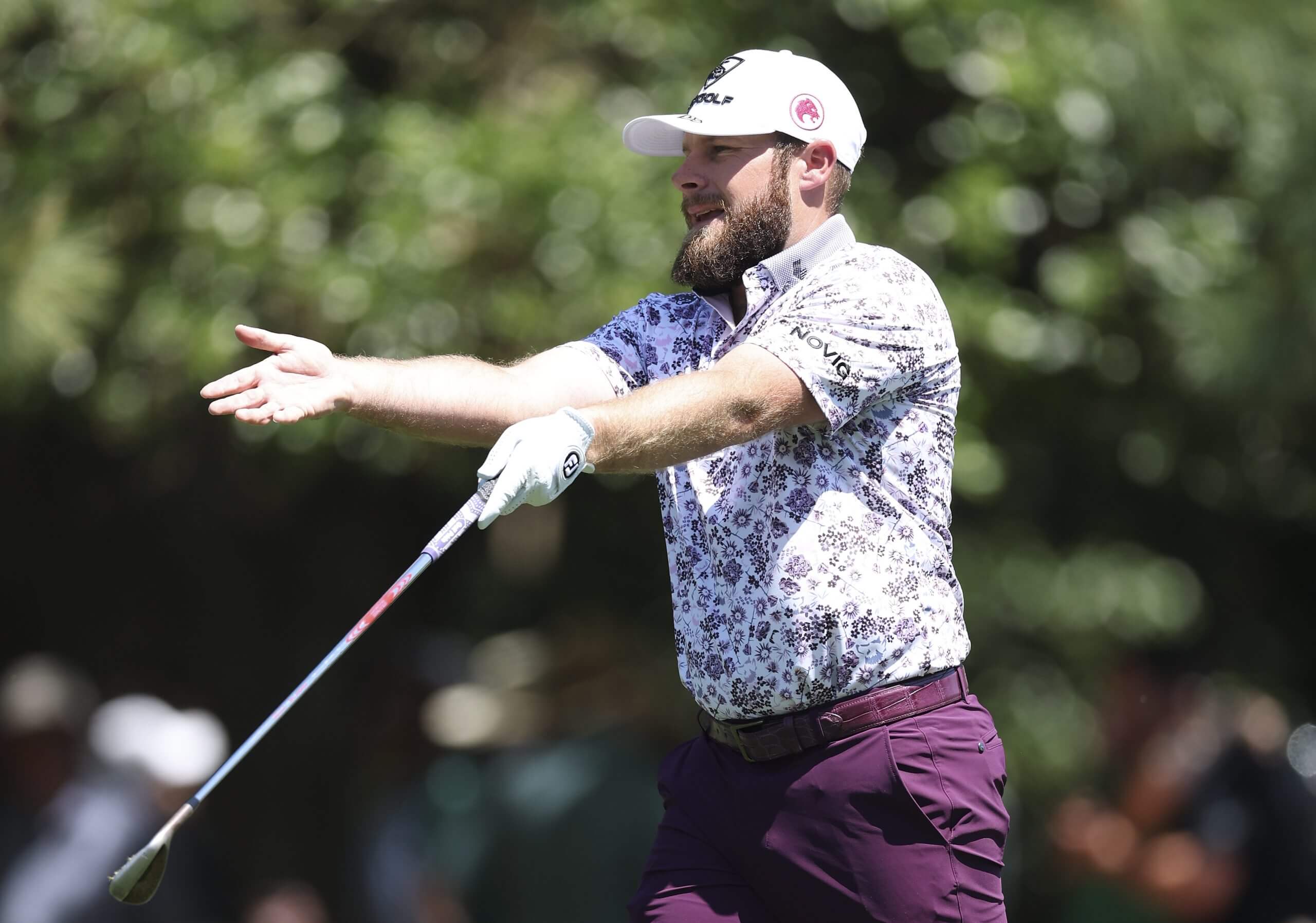 Tyrrell Hatton, in purple pants and a purple and white floral-patterned shirt and white ball cap, holds his club with his left hand and gestures with his right. 