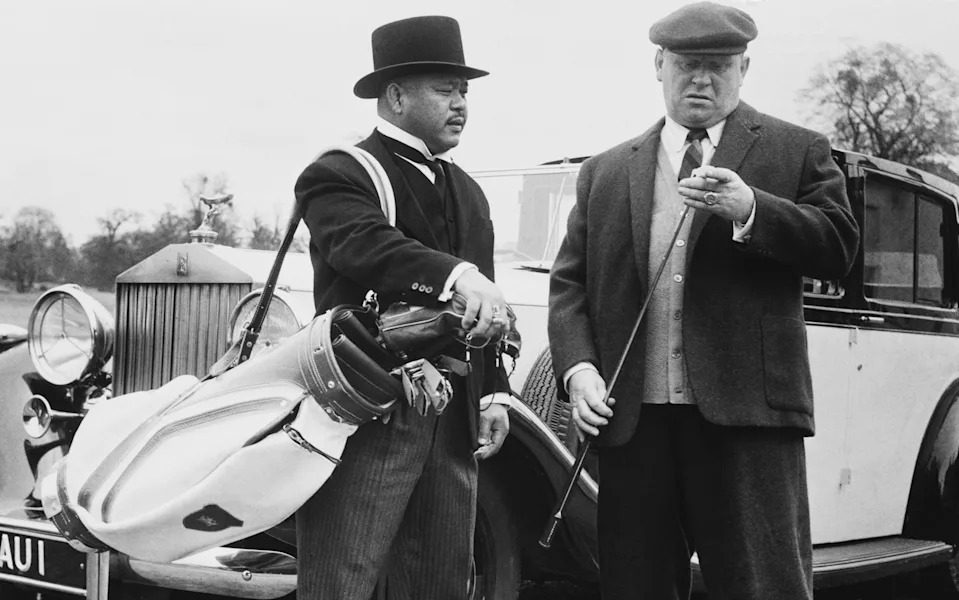 Auric Goldfinger looks over his club as his assistant Oddjob holds his clubs in Goldfinger